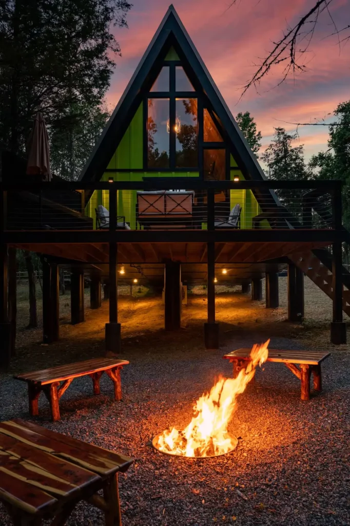 Striking green A-frame cabin elevated over a fire pit area at dusk