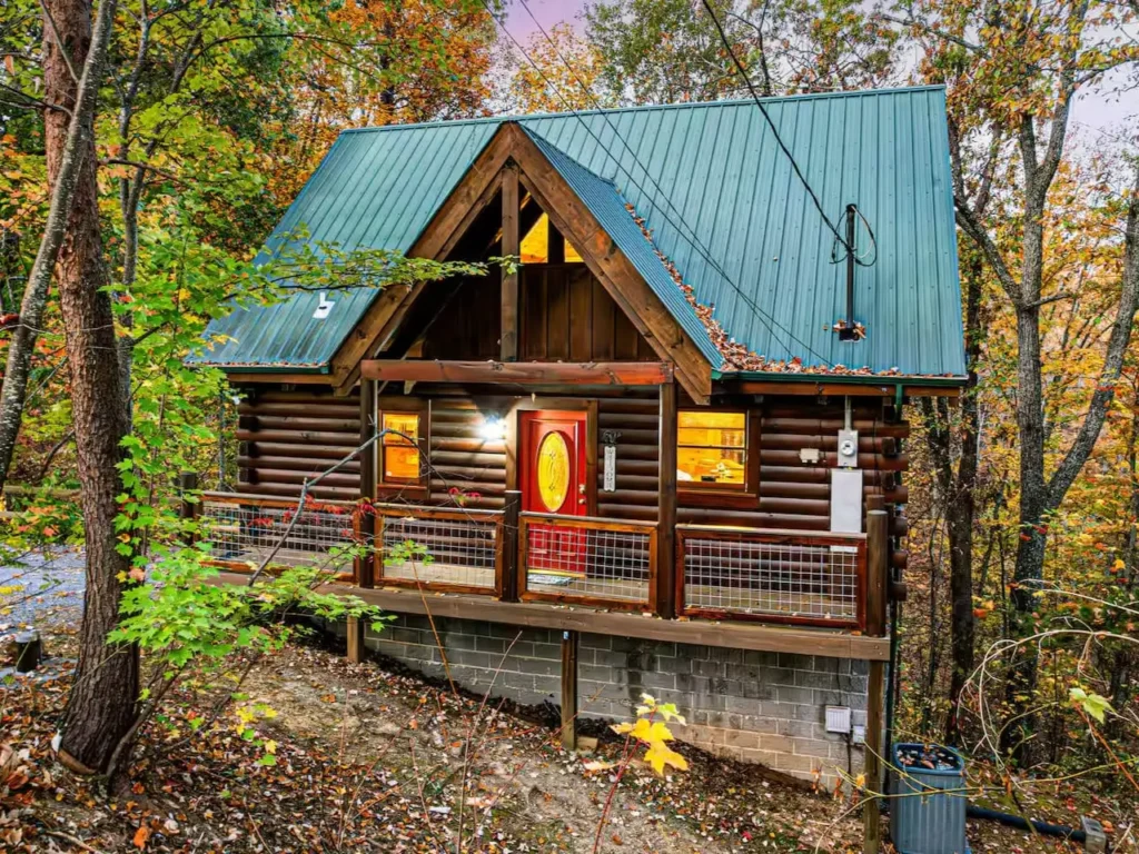 Log cabin with green metal roof and red door, nestled on a slope in autumn