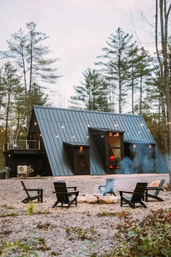 Modern Maine A-frame cabin exterior at dusk with a cozy fire pit area and string lights in a wooded setting.