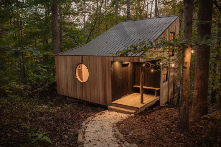 The Nook Cabin in Swannanoa, NC, a quaint wooden tiny home with a glowing round window, nestled in a twilight forest setting.