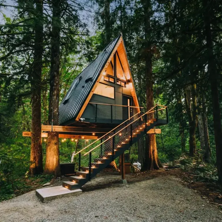 Modern elevated A-frame Treeframe Cabin rental glowing amidst tall forest trees in Index, Washington