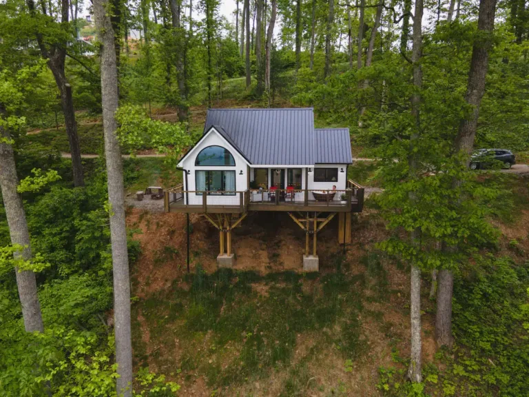 Elevated white modern farmhouse tiny home, 'The Farmhouse - Earth & Sky Dwellings,' surrounded by lush green trees on a hillside.