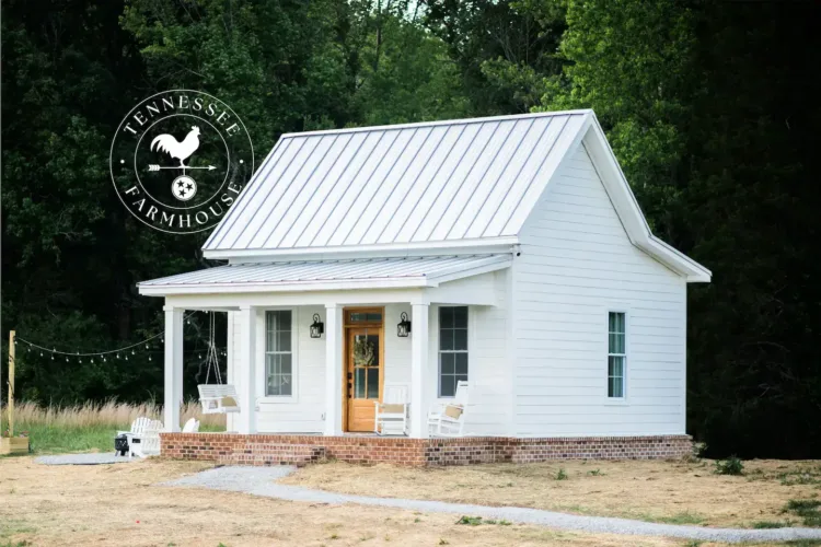 Classic white Harriman Tiny Farmhouse in Tennessee, featuring a covered porch with a swing, set against a backdrop of trees under a clear sky.