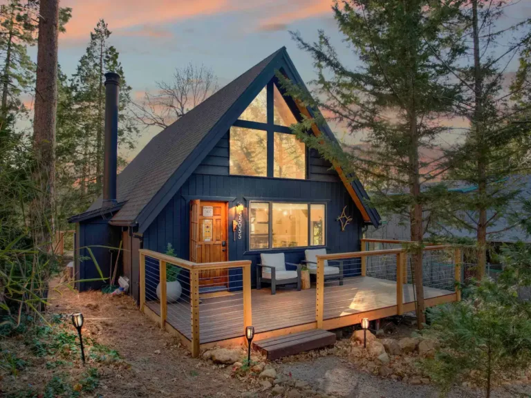 Striking Wildwood A-frame Cabin in Idyllwild, California, illuminated at dusk, with a large wooden deck and expansive windows, surrounded by tall pines.