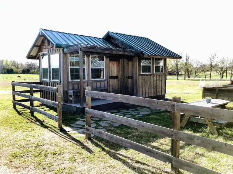 Rustic Mars Hill Farm Tiny House in Ferris, Texas, a wooden cottage with a green metal roof and a fenced yard with a picnic table, on a sunny day.