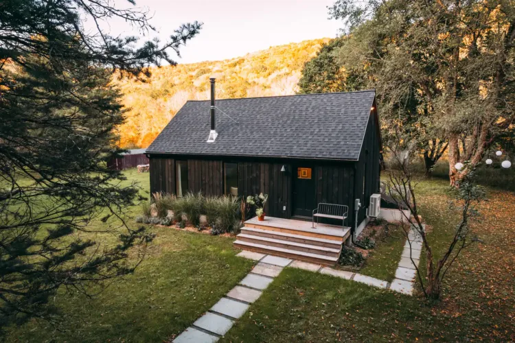 Dry Brook Cabin, a dark modern tiny home in Arkville, NY, with clean lines and landscaped entry, set in a grassy yard with autumnal trees.