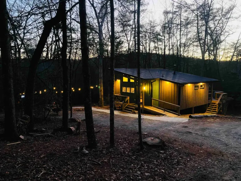 Trahlyta modern cabin in Dahlonega, GA, illuminated at dusk in a wooded setting, with string lights adding a warm glow to the surroundings.