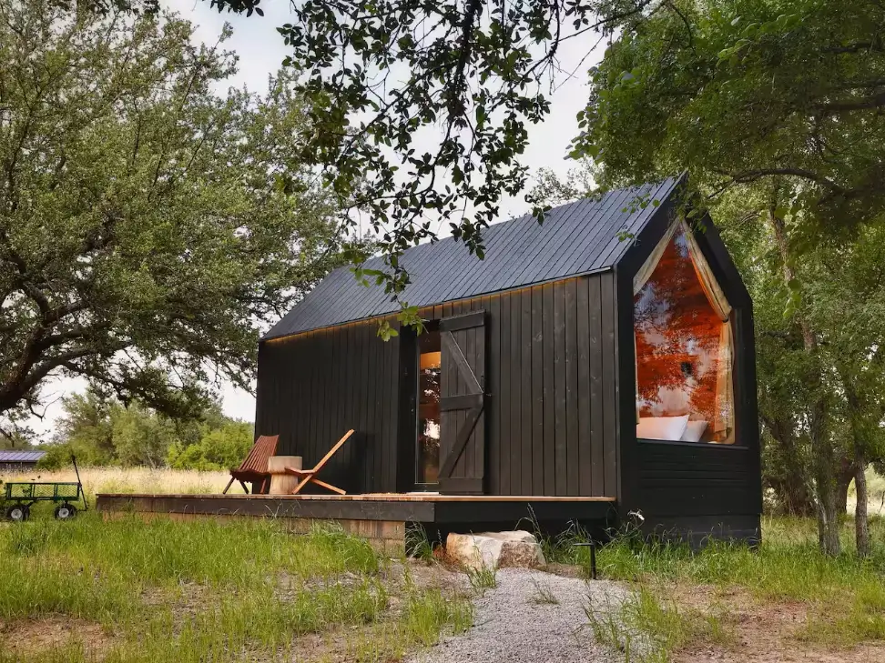 Sleek black Now Here Cabin with a large triangular window and a simple wooden deck with two chairs, set in a tranquil natural environment.