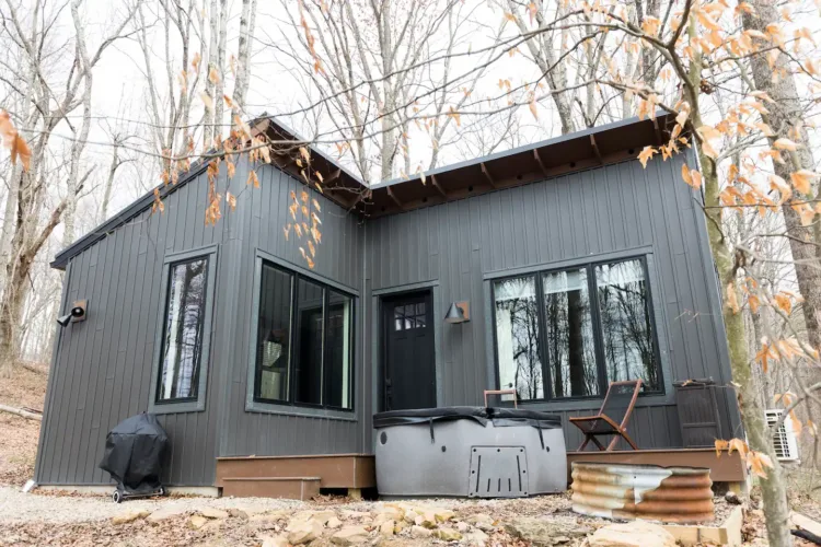 Aspen Haus, a modern grey tiny cabin in New Plymouth, OH, with a covered hot tub on its deck, set in a leafless woodland during cooler weather.