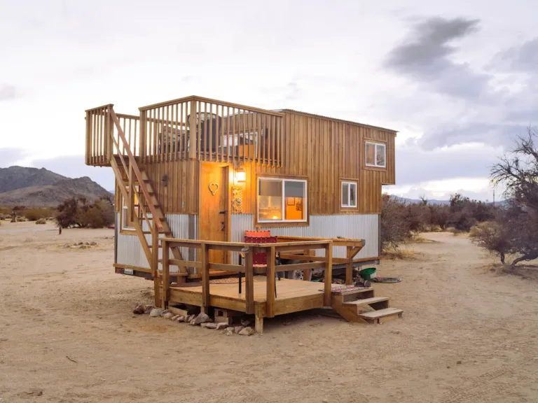 Peacock Tiny House in Sandy Valley, NV, a wooden tiny home with corrugated metal accents, featuring a lower deck and an upper rooftop deck, set in a desert landscape at dusk.