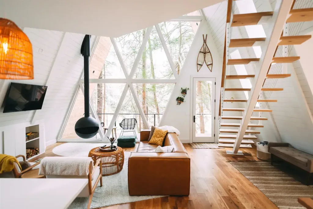 Bright A-frame living room with white walls, high ceiling, modern black hanging fireplace, leather sofa, and large triangular window showing trees.