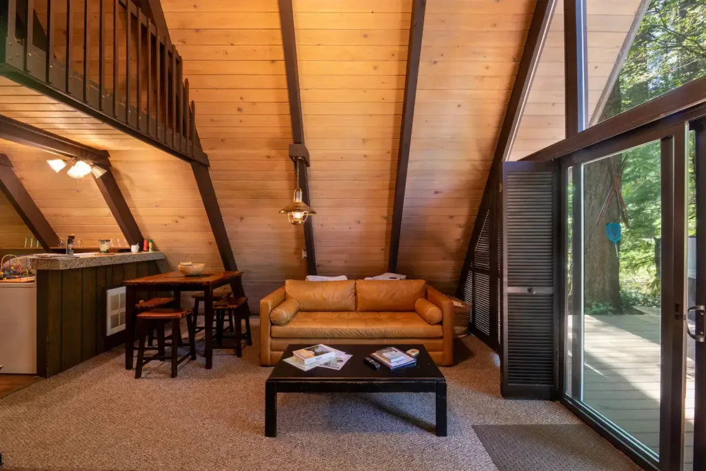 Cozy A-frame living room with full wood paneling, tan leather sofa, small dining set, and sliding doors leading to an outdoor deck.
