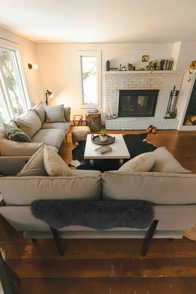 Bright living room with a white painted brick fireplace, large light gray sectional sofa, hardwood floors, and a faux fur throw.