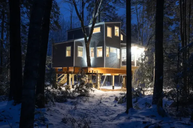 Modern multi-story treehouse rental in Hancock Maine, lit up at night in a snowy forest setting, offering a luxurious winter escape.