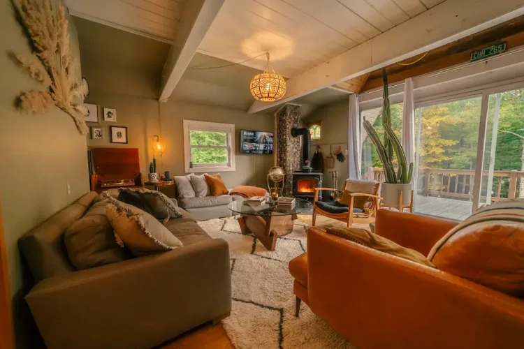 Eclectic A-frame living room with sage green walls, a mix of comfortable sofas, a patterned rug, a wood stove, and various personal decorative items.
