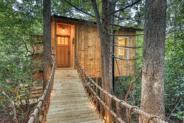Charming wooden treehouse rental in Old Fort North Carolina, accessed by a rope suspension bridge with a welcoming lit doorway.