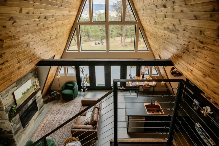 High-angle view of an A-frame interior with a wood-paneled ceiling, large triangular window, open living and dining area with a stone fireplace, and a loft railing.