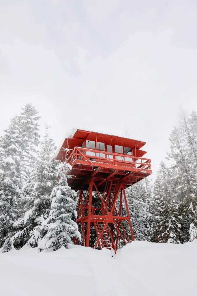 Bright red Crystal Peak Lookout fire tower treehouse rental in Fernwood Idaho, surrounded by snow-covered evergreen trees.