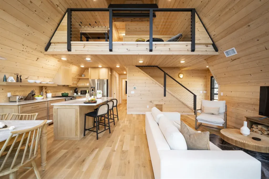 Bright modern A-frame interior with extensive light wood paneling, a white sofa, a kitchen with an island, and a loft space visible above with black railings.