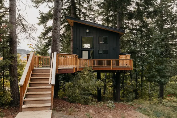 Modern black Klickitat Treehouse rental in White Salmon Washington with an angled roof and spacious light wood deck among trees.