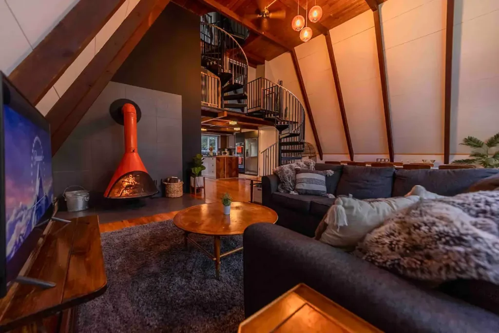 A-frame living room featuring a prominent orange retro freestanding fireplace, a black spiral staircase, a dark gray sofa, and a wood-paneled ceiling.