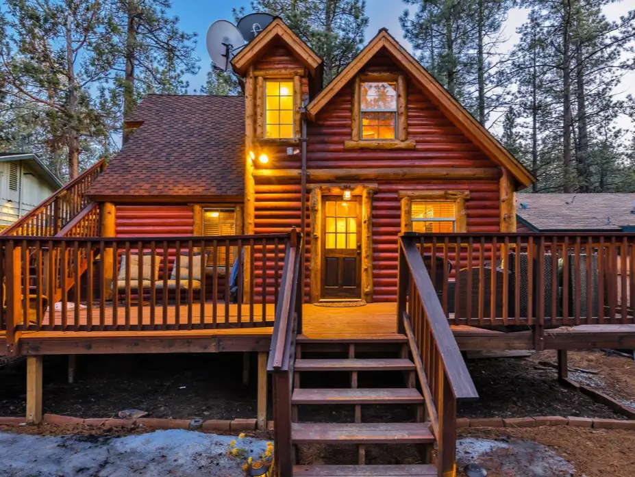 An inviting two-story Big Bear log cabin glows warmly at dusk, surrounded by pine trees.