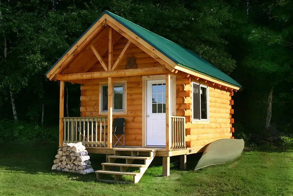 The Little Log Cabin kit with a green metal roof, set in a summer landscape with a canoe and firewood.