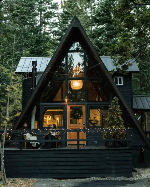 The stylish, dark exterior of the Mid-Century Modern Tahoe A-frame cabin in Homewood, illuminated by warm interior lights and surrounded by tall pine trees at dusk.