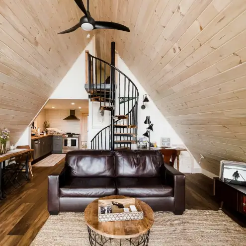 A-frame living area with a very high, light wood-paneled ceiling, a brown leather sofa, a round woven coffee table on a jute rug, and a black spiral staircase.