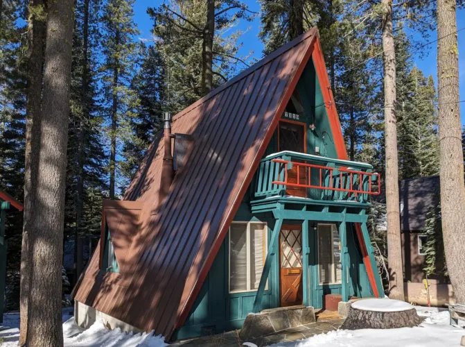 The Chickaree Mountain Retreat, a vintage Tahoe A-frame cabin in Soda Springs, featuring a distinctive teal and red color scheme, pictured on a sunny, snowy day among the pines.