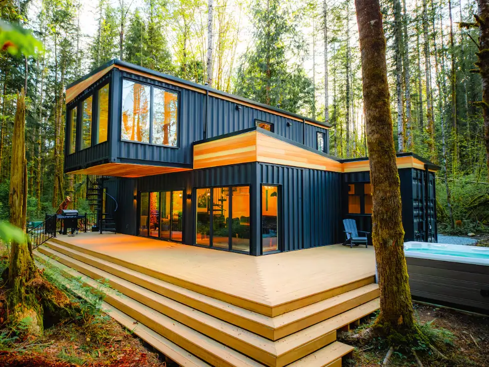 Design of The Pacific Bin, a modern shipping container home in Monroe, WA, highlighting a dark exterior with wood details, a multi-tiered deck, hot tub, and forest integration.