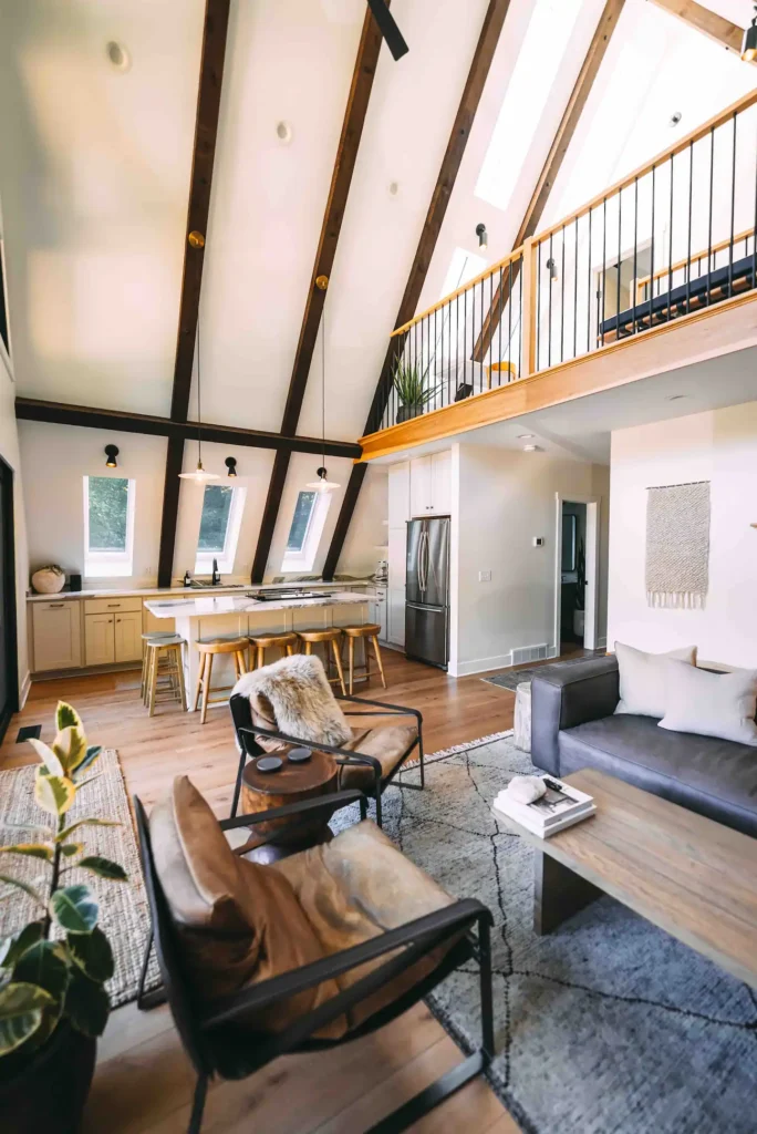 Modern A-frame interior with white walls, dark exposed ceiling beams, skylights, a contemporary kitchen with an island, and a visible loft area.