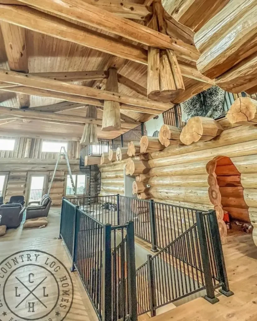 The grand interior of a handcrafted log home featuring a vaulted ceiling with massive timber trusses and a loft with wrought iron railings.