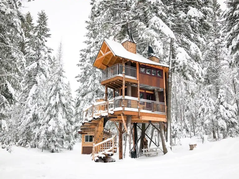 Multi-story wooden Meadowlark Treehouse at Montana Treehouse Retreat, covered in snow, with a unique spiral staircase around a fir tree.