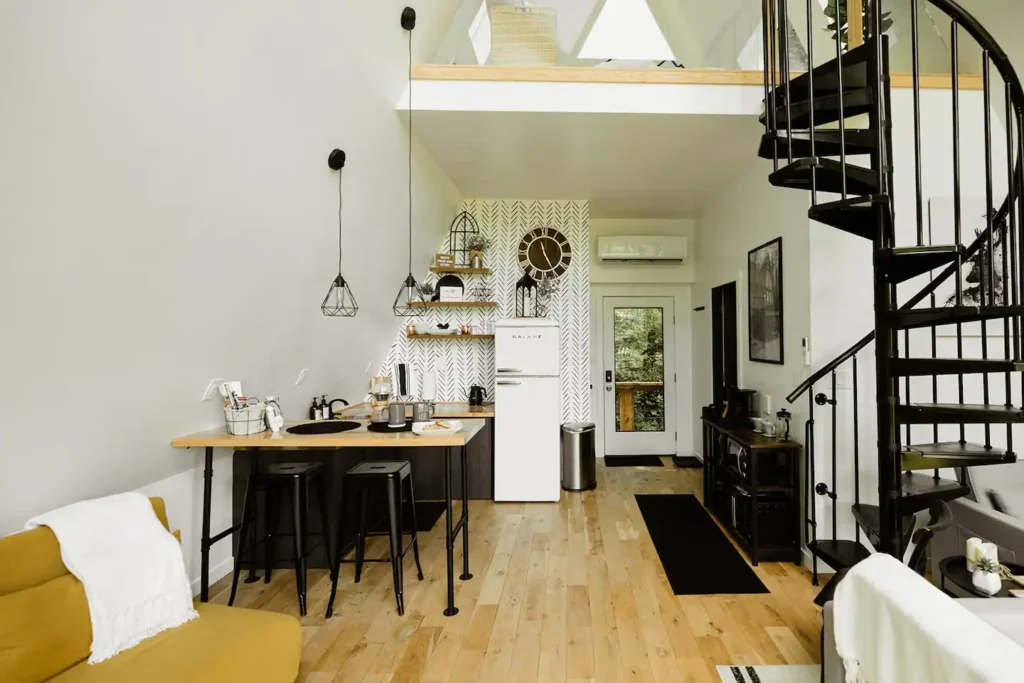 Stylish compact A-frame with a kitchenette featuring a herringbone accent wall, a black spiral staircase, light wood floors, and a yellow armchair.