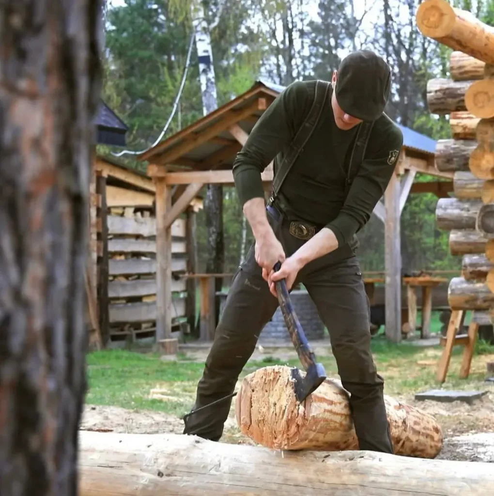 A craftsman in suspenders uses a traditional axe to hand-shape a notch in a large log.