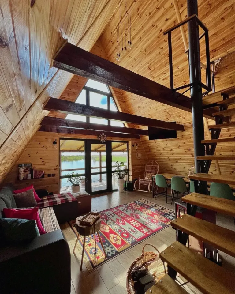 Warm wood A-frame house interior ('Tarska carolija') featuring a spiral staircase, lake view, and colorful rug.