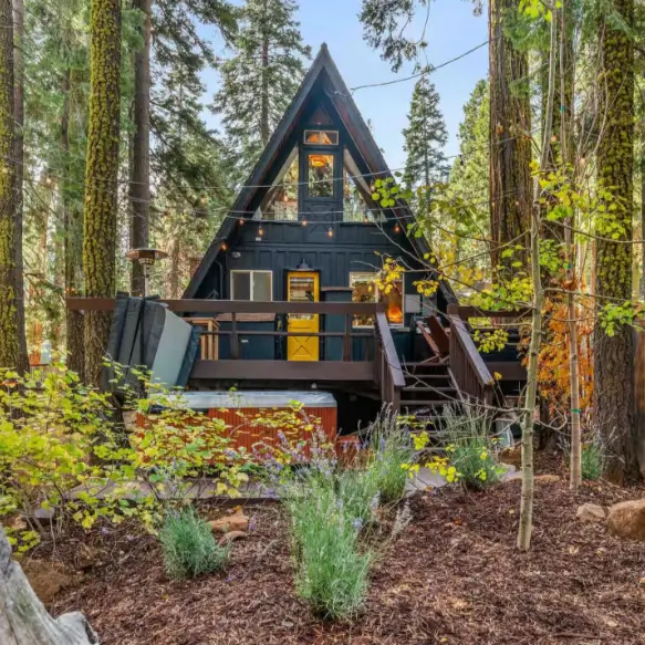 The Sugar Pine Speakeasy, a cozy Tahoe A-frame cabin in Homewood, featuring a hot tub on its deck and a distinctive yellow door, nestled in a lush green forest.
