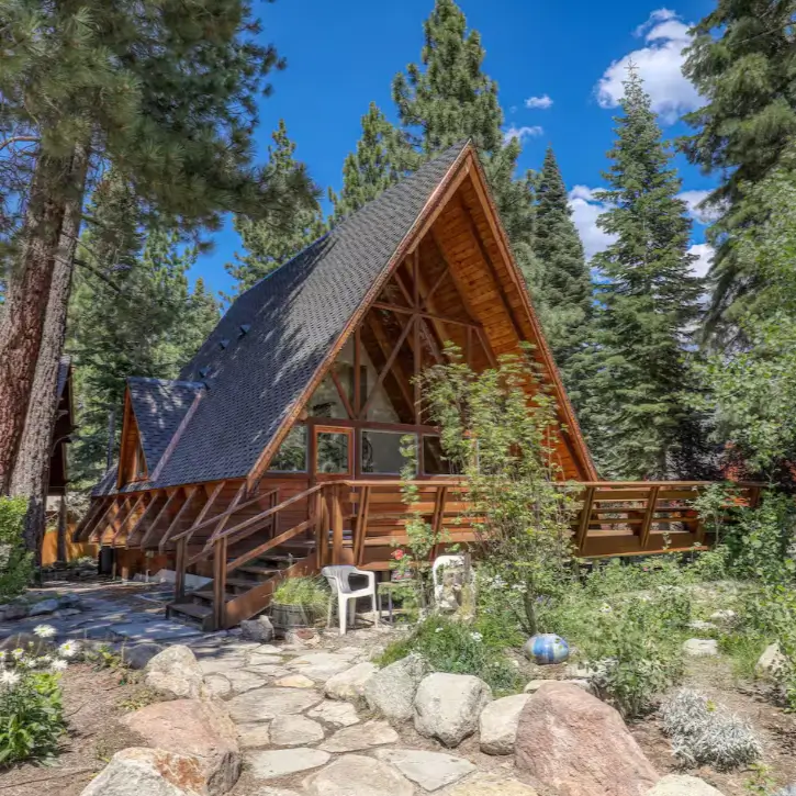 A classic, wood-sided Tahoe A-frame cabin in Carnelian Bay, surrounded by a lush garden and tall pine trees on a bright, sunny day.