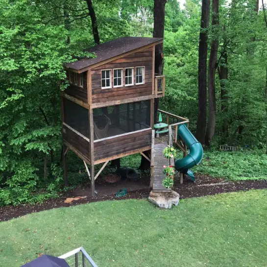 Two-level wooden Trailside Treehouse rental in Richmond Virginia, with a screened-in porch and a green tube slide, bordering a lawn and woods.