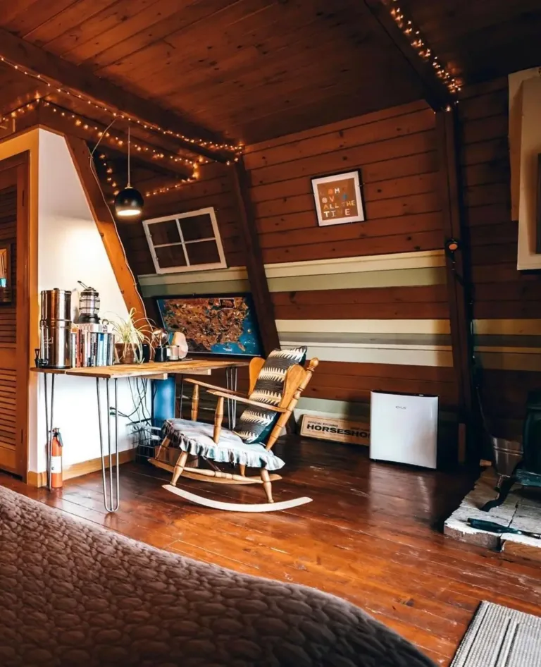 Cozy small A-frame cabin interior with wood paneling, rocking chair, desk nook, and warm string lights.