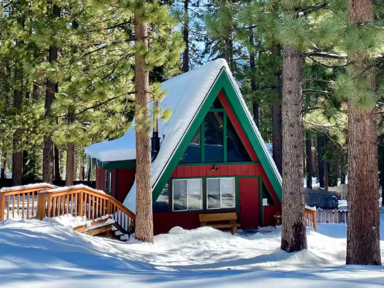 The Westcabinn, a classic red Tahoe A-frame cabin in South Lake Tahoe, shown with its roof covered in snow on a sunny winter day among the pines.