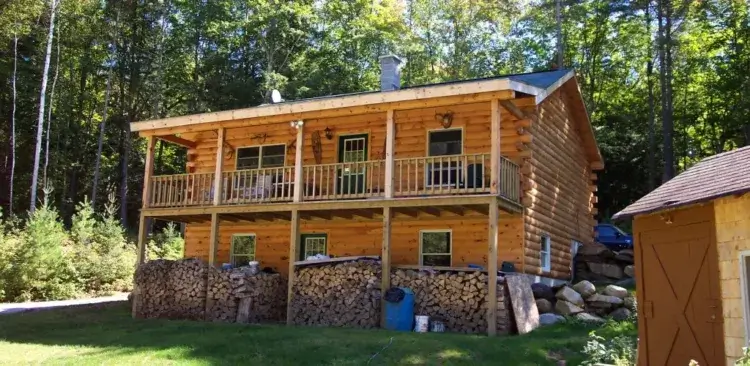 The two-level Adventurer log cabin on a sloped property with large stacks of firewood under the porch.