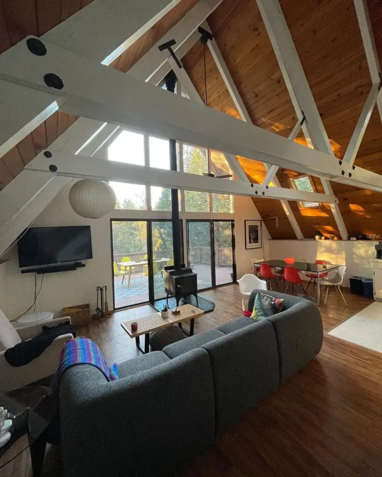 Bright simple A-frame house interior with white structural beams, wood ceiling, modern stove, and large windows.