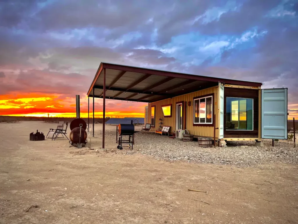 El Coyote, an off-grid shipping container home design in Terlingua, TX, featuring a simple tan exterior and covered patio, perfectly framing a dramatic desert sunset.