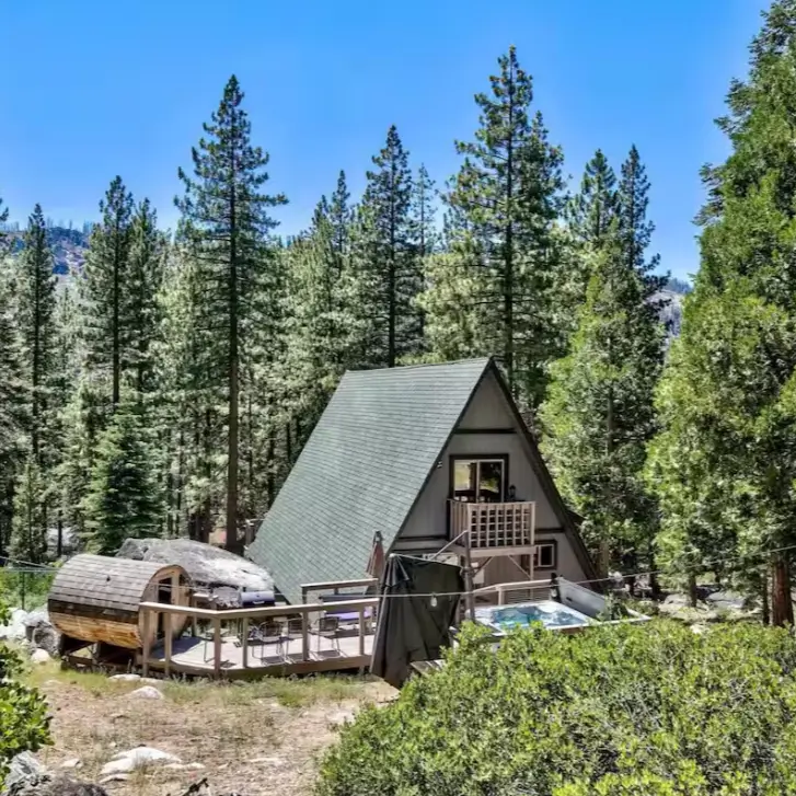 The secluded Reindeer Cabin, a classic Tahoe A-frame in South Lake Tahoe, featuring a deck with a hot tub and barrel sauna nestled in the forest.