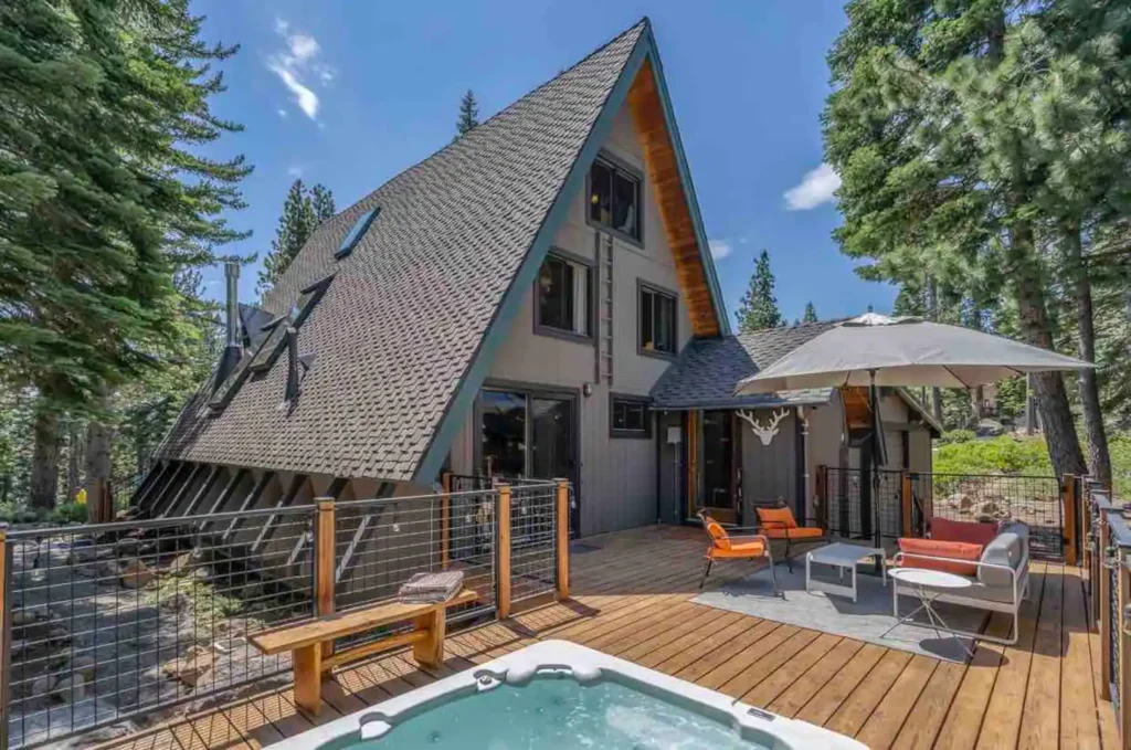The spacious deck of an updated West Shore Tahoe A-frame cabin in Homewood, featuring a hot tub and comfortable patio furniture under a sunny sky.