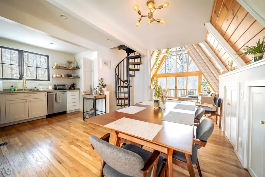 Bright open-concept A-frame showing a dining table in the foreground, a kitchen to the left, a black spiral staircase, and a living area with large windows in the background.