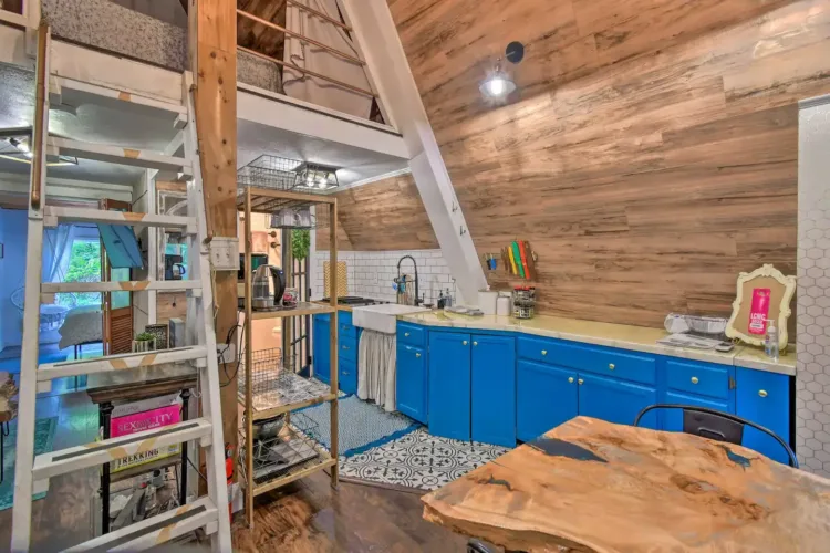 Eclectic A-frame barn loft studio interior featuring bright blue kitchen cabinets and a ladder to the loft.
