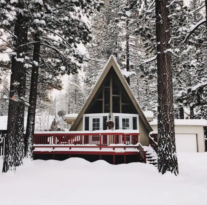 A classic Tahoe A-frame chalet in South Lake Tahoe, covered in a thick blanket of snow, with a red-railed deck surrounded by tall, snow-dusted pine trees.